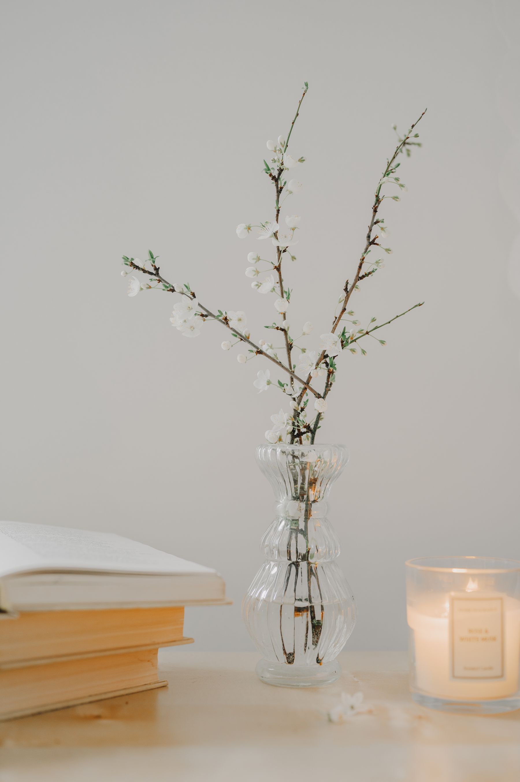 White blossoming branches in a glass vase beside stacked books and a candle